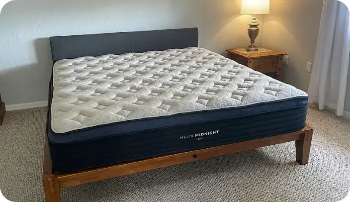 A mattress on a wooden bed frame in a bedroom setting, with a bedside lamp and wall visible in the background. The mattress has a quilted light beige top and a dark navy blue side panel with the brand name 'HELIX MIDNIGHT' visible.