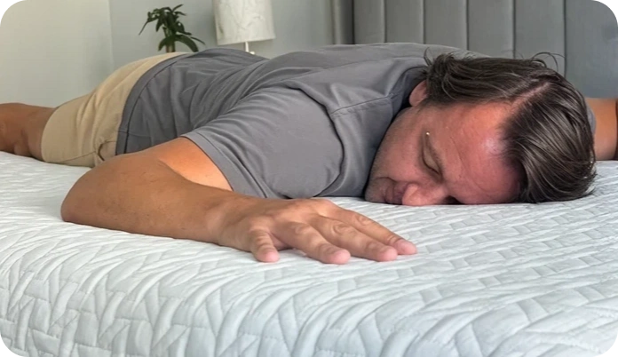 A man lying face down on a white quilted bedspread in a bedroom setting, wearing a gray t-shirt and beige shorts.