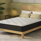 A mattress on a light wood bed frame with a tall matching headboard, styled in a bedroom setting with pillows, a side table, lamp, and plant.